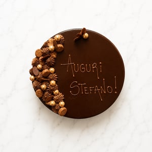 Torta al cioccolato decorata con scritta “Auguri Stefano!”, macarons, ciuffi di crema e dettagli dorati su sfondo marmo bianco.