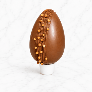 Uovo di Pasqua in cioccolato al latte con decorazioni verticali e sfere croccanti, esposto su supporto bianco minimal.