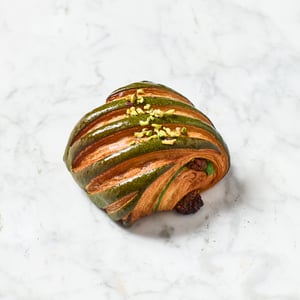 Viennoiserie artigianale con sfoglia bicolore al pistacchio, ripiena e decorata con granella di pistacchio, fotografata su una superficie in marmo chiaro